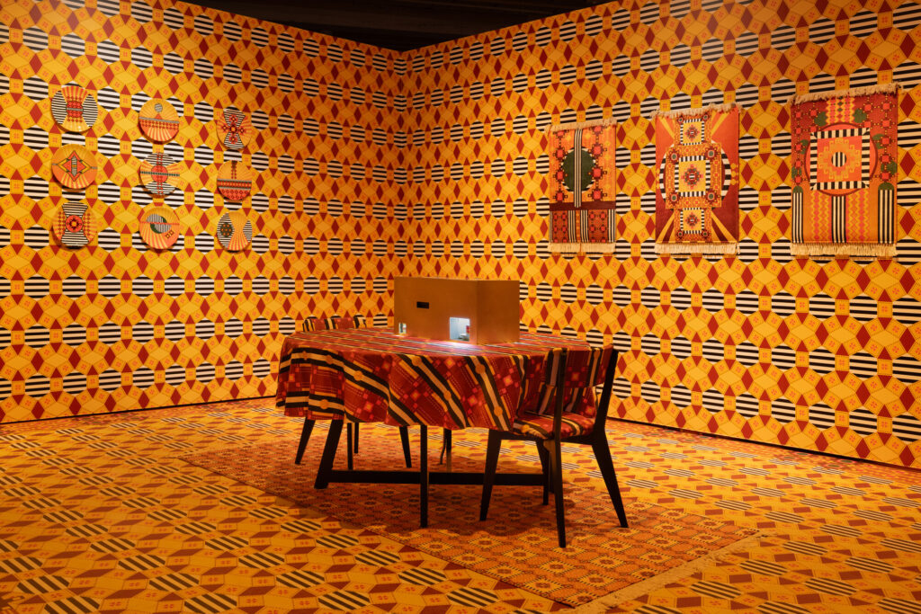 Installation photo featuring AI-assisted tasseography readings, brass enclosure, Armenian patterns on wall paper, flooring, textiles and ceramics, with furniture, and books.
